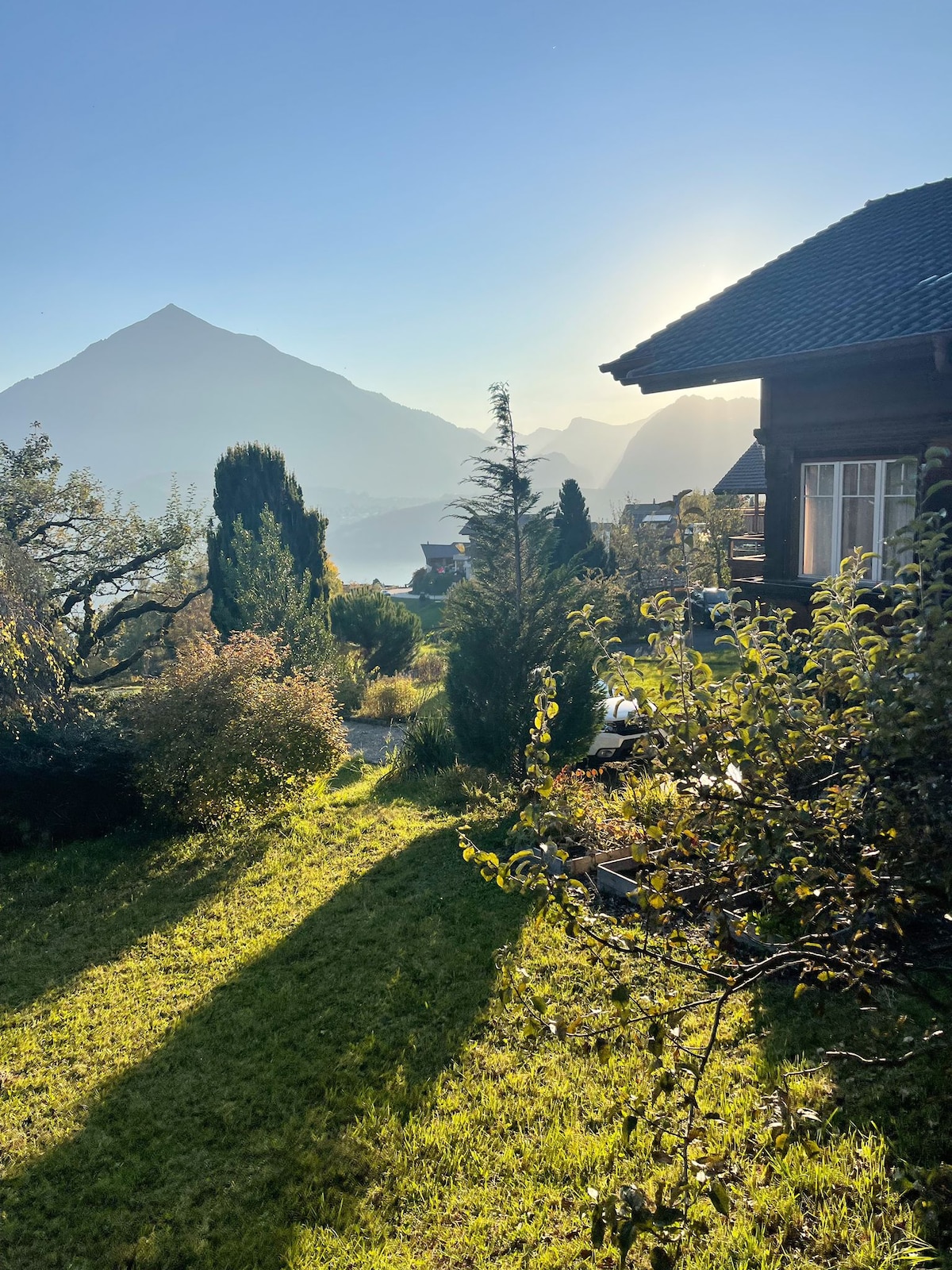 A scenic view of the chalet's exterior is presented, showcasing lush greenery and well-maintained garden areas. The backdrop features majestic mountains with the sun rising, creating a serene atmosphere. Soft shadows cast by trees add depth to the landscape, enhancing the natural beauty.