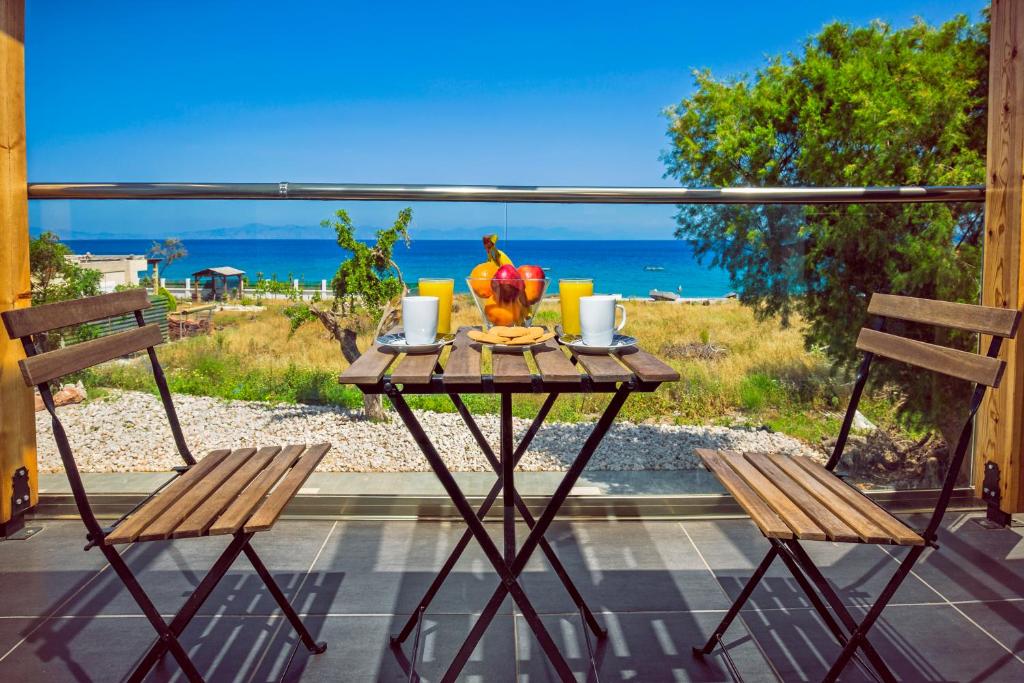 A small outdoor dining area features a wooden table with two folding chairs, set against a backdrop of a clear blue sea. Fresh fruit is arranged on the table, accompanied by two cups. Lush greenery is visible in the distance.