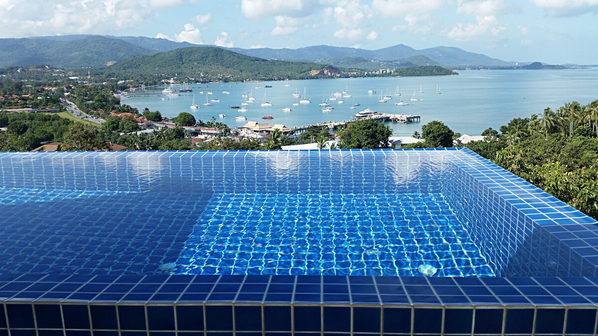 An infinity pool features vibrant blue tiles, overlooking the bay with views of the water and distant hills. Sailboats can be seen in the harbor, while a village is nestled along the shoreline, creating a serene coastal landscape.
