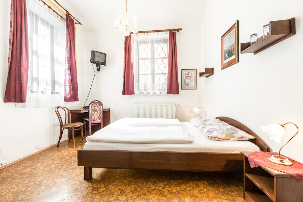 A bright room features a spacious bed with decorative pillows, accompanied by bedside lamps and a small table. A desk with a chair is positioned near the window, which is adorned with sheer curtains and red drapes, allowing natural light to enter the space.