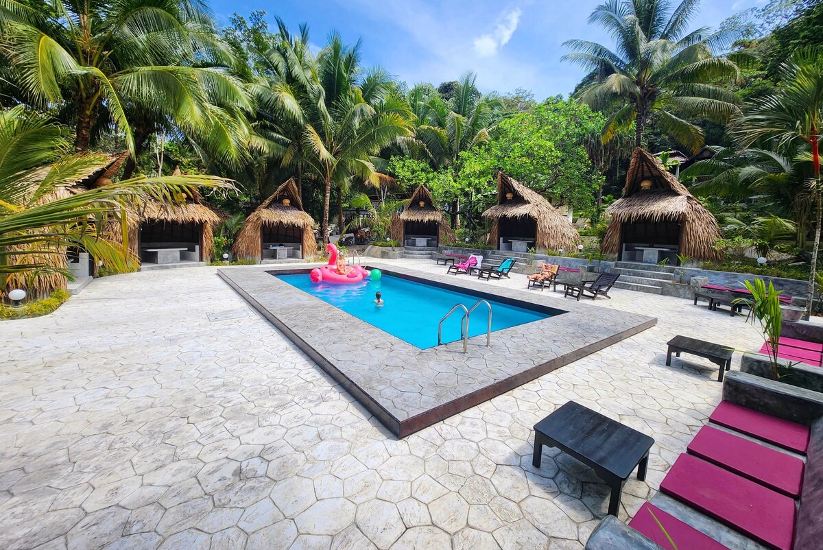 A swimming pool is surrounded by tropical palm trees and thatched-roof bungalows. Sun loungers and tables are arranged on a stone patio, providing ample lounging space. Colorful pool floats add a playful touch to the water, inviting relaxation and enjoyment in the lush setting.