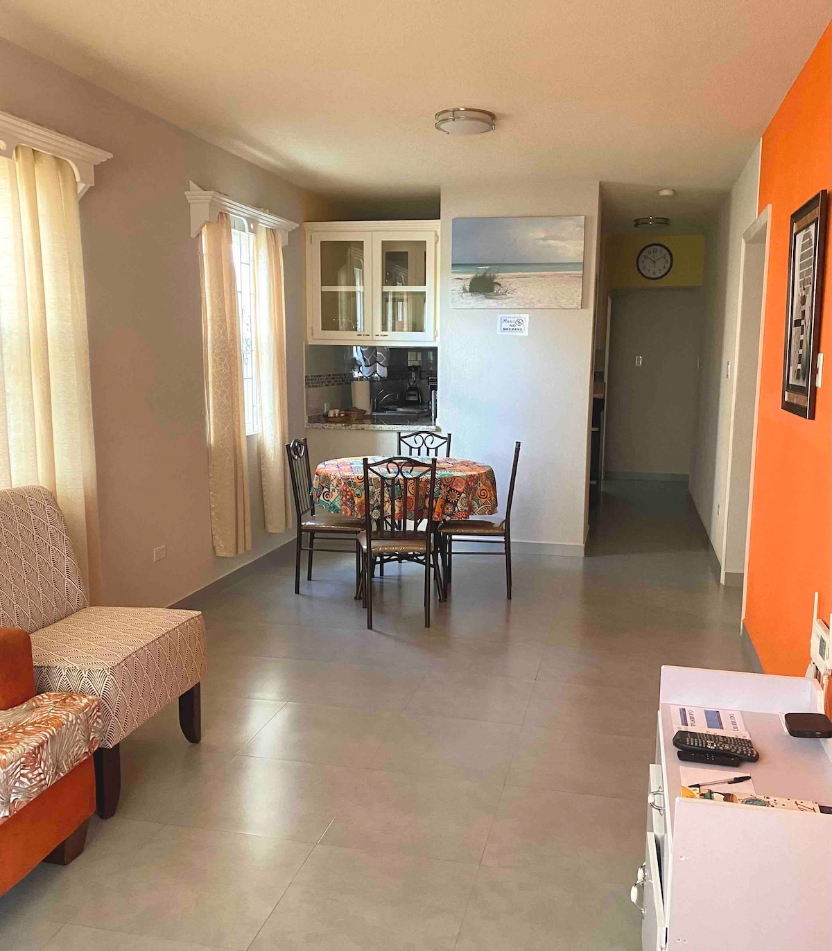 A living area features a patterned dining table surrounded by four black chairs, with sunlight filtering through sheer curtains. An orange accent wall provides contrast to the neutral tones, while a comfortable chair and a side table are positioned near the window. A kitchen area is visible in the background.