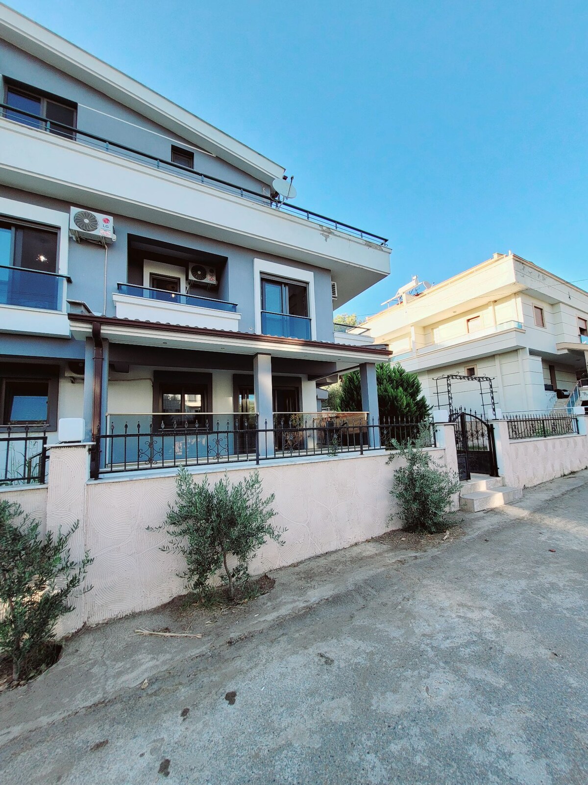 A three-story villa is presented with a modern exterior, featuring a stone wall and a patio area. The entrance is framed by shrubs, and air conditioning units are visible on the facade. A clear blue sky creates a bright backdrop.
