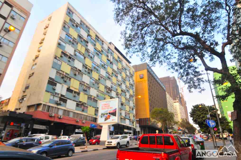 A multi-story apartment building is positioned next to modern structures along a busy street. Windows are visible with varying shades of pane coverings, and parked cars line the street. Trees provide some greenery, contrasting the urban setting.