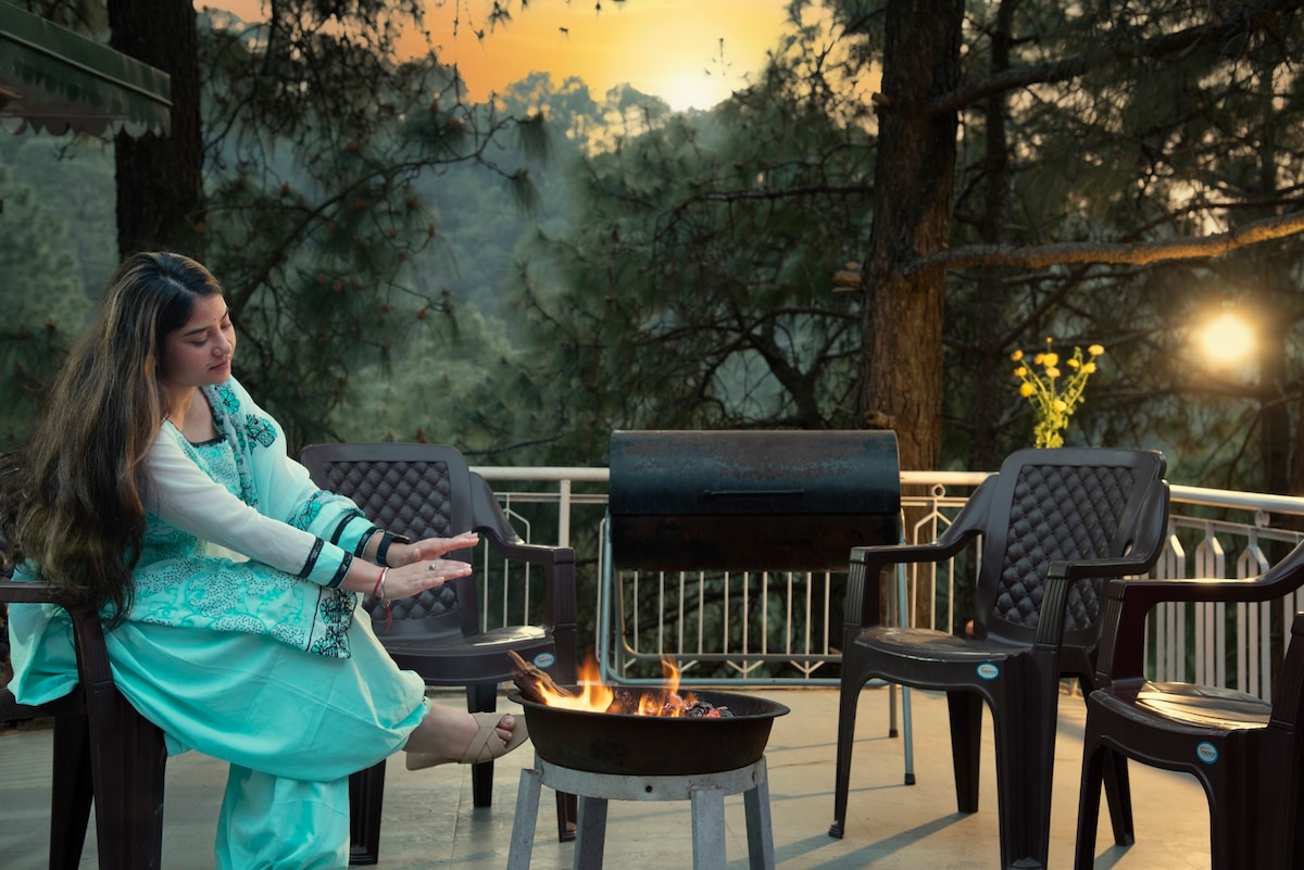 An outdoor seating area is highlighted by a fire pit surrounded by several chairs. Soft sunlight filters through the trees in the background, creating a warm ambiance. The space is designed for relaxation and social gatherings, offering a serene view of the surrounding nature.