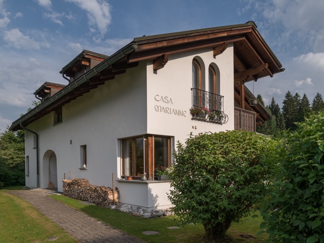 The exterior of a two-story home is presented, featuring a white façade with wooden accents. A balcony with flower boxes overlooks a grassy area. Logs are stacked neatly beside the entrance, and the surrounding landscape is green, enhancing the overall serene ambiance.