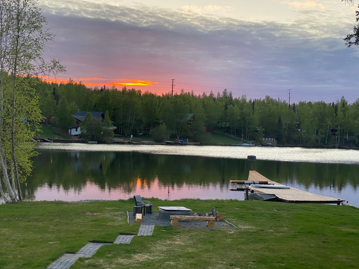 The image captures a serene lake view during sunset, with soft pink and purple hues reflected in the water. A grassy area leads to a wooden dock, with seating options and a fire pit nearby. Trees line the opposite shore, enhancing the landscape.
