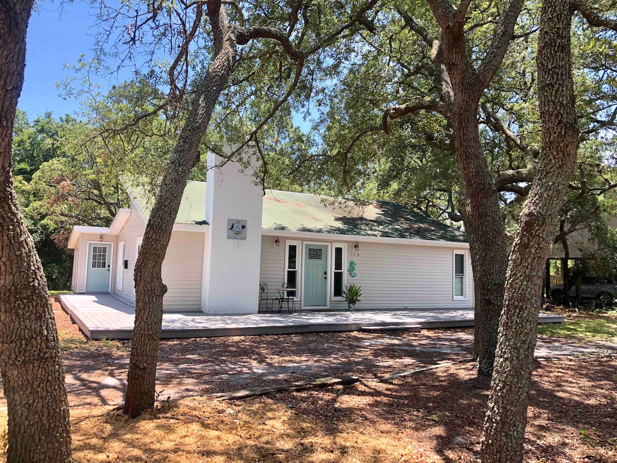 The home is framed by mature trees, providing shade and privacy. A covered porch welcomes guests at the entrance, with a seating area visible. The exterior features a light-colored facade and a simple, pitched roof, set on a spacious lot surrounded by natural elements.