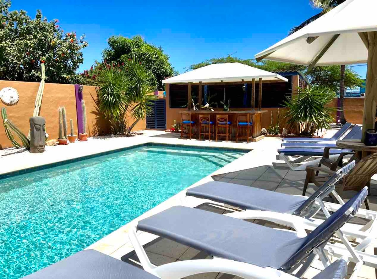 A clear blue pool is surrounded by lounge chairs and tropical greenery, providing a tranquil outdoor space. A shaded dining area with high chairs is situated near the pool, complemented by a gazebo and various plants, enhancing the relaxing atmosphere.