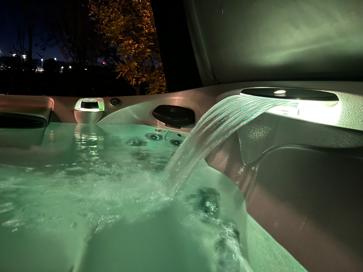 A hot tub is shown with a gentle water feature cascading into the warm water. Multi-coloured lighting illuminates the tub, creating a calming ambiance. The surrounding area reflects a serene night setting with hints of nature visible in the background.