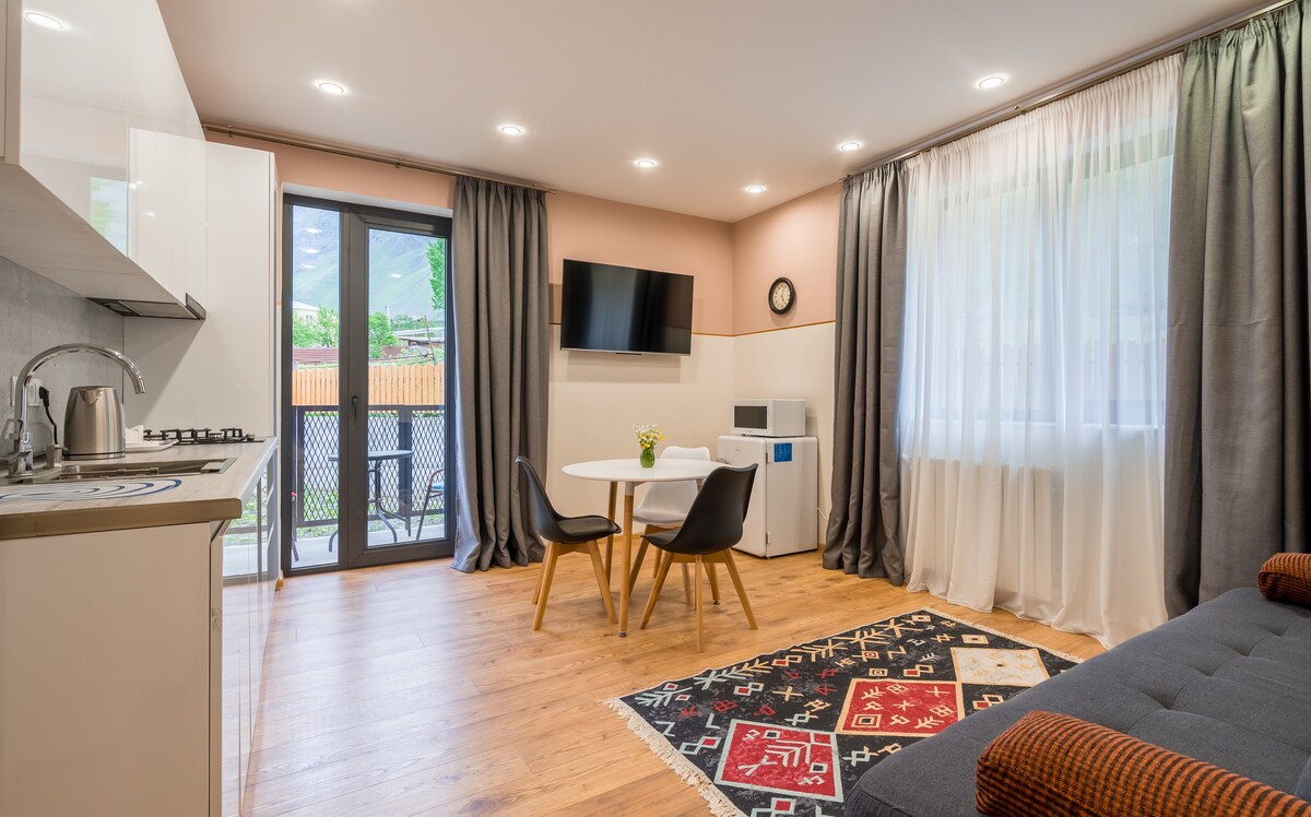 A modern living space includes a kitchenette with sleek cabinetry and a dining area featuring a round table with two chairs. A television is mounted on the wall, and light enters through large windows adorned with sheer curtains, creating a bright atmosphere. A balcony door opens to outdoor views.