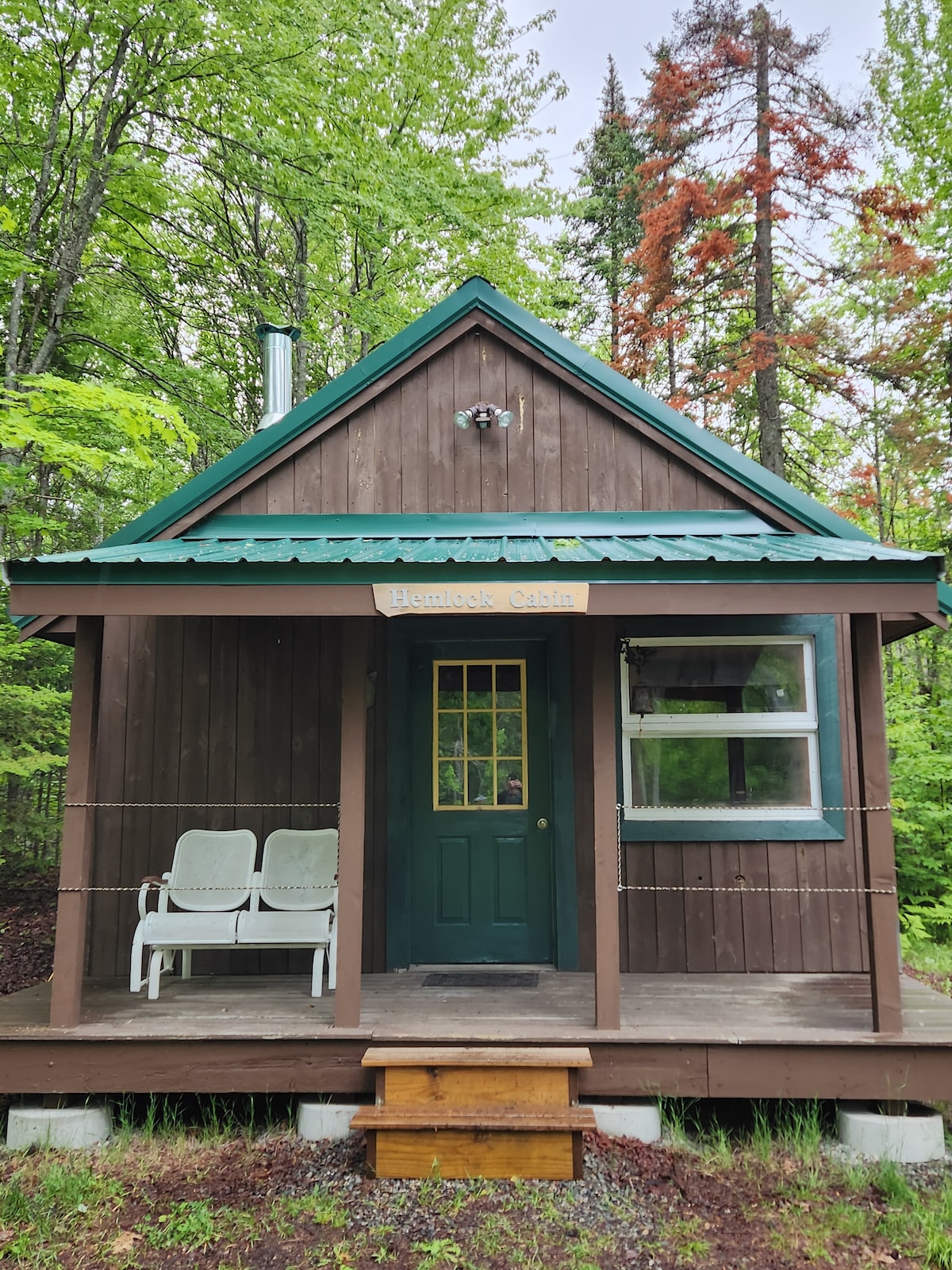 The Hemlock Cabin features a rustic wooden exterior with a green metal roof and an inviting front porch. Two white chairs are positioned on the porch, and a green door with a window adds charm. Surrounding trees create a natural setting.