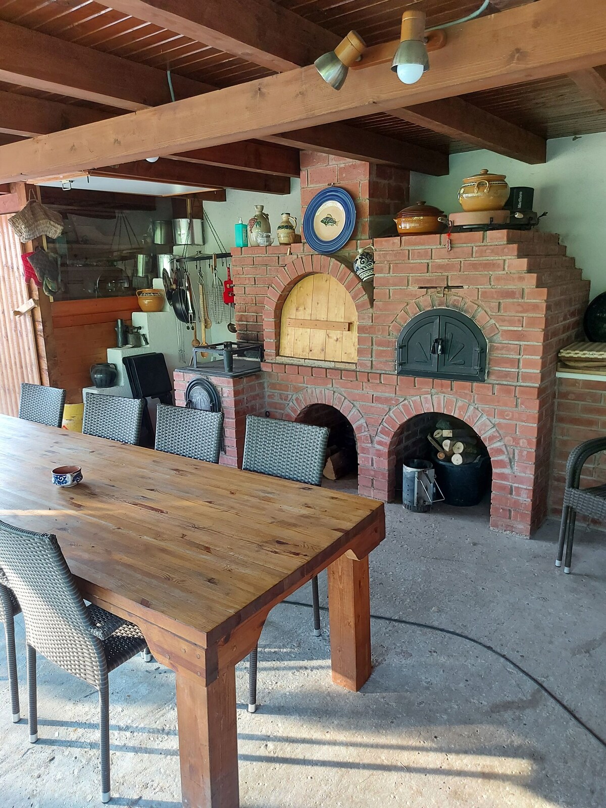 A rustic dining area features a large wooden table surrounded by several black chairs. A traditional brick oven and fireplace are positioned against the wall, adorned with various pots and decorative items. Natural light illuminates the space, enhancing the warm wood tones.