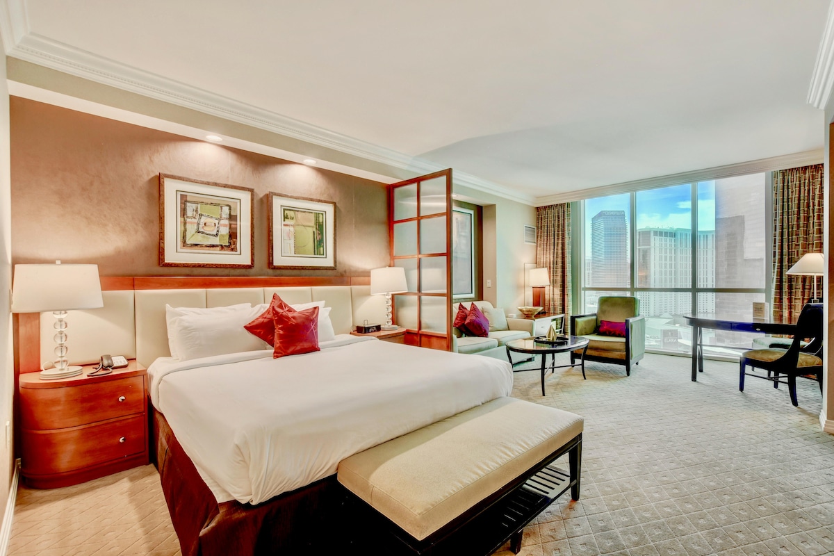 A spacious suite is presented featuring a king-size bed with crisp linens and accent pillows. Large windows bring in natural light, offering views of the cityscape. Comfortable seating options and a desk are thoughtfully arranged to enhance the room's functionality.