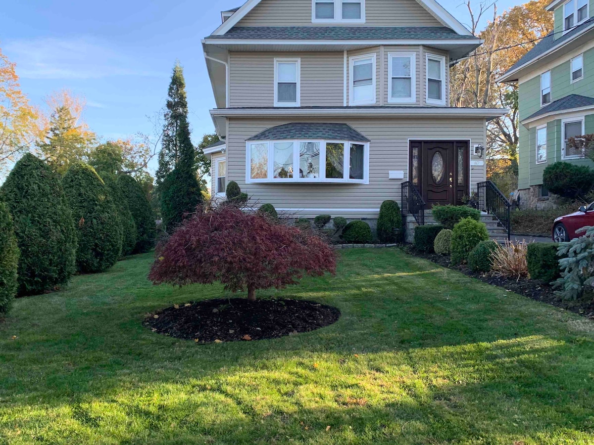 A multi-story house is showcased, featuring a welcoming entryway with a dark wooden door. The front yard is landscaped with neatly trimmed bushes and a small flowering tree, surrounded by a well-maintained lawn that extends towards the sidewalk.