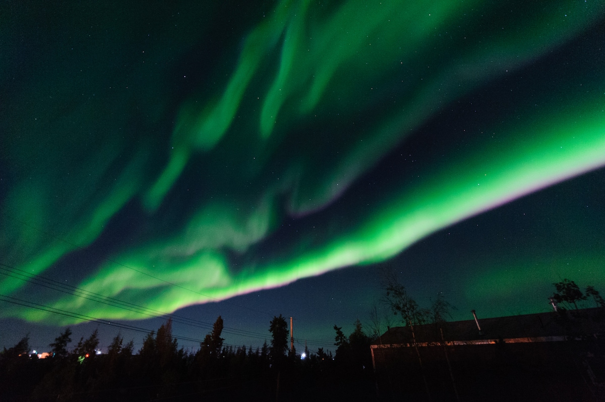 The night sky is illuminated by vibrant green northern lights, casting a captivating glow over the landscape. Dark silhouettes of trees and buildings are visible beneath the colorful streaks, enhancing the tranquil nighttime setting.