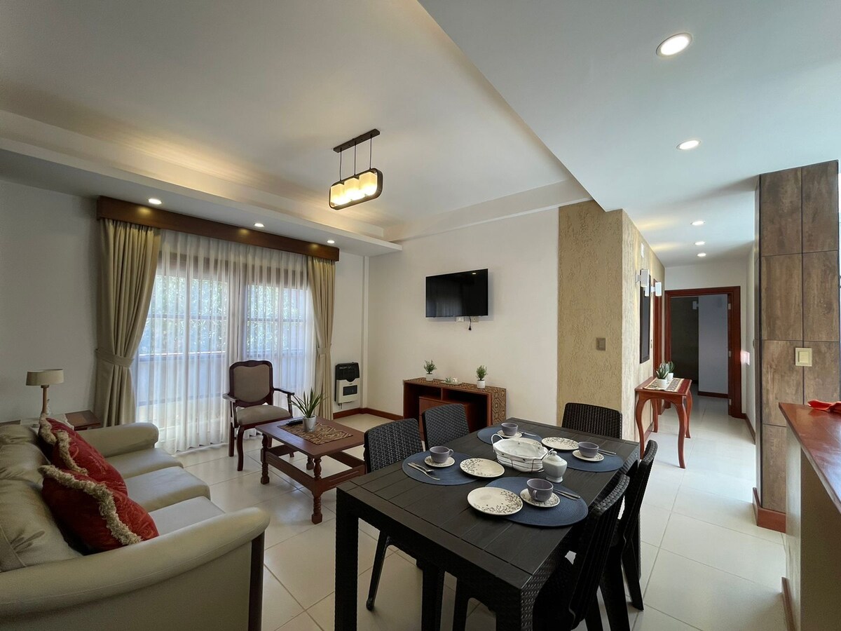 A bright living area is depicted, featuring a comfortable sofa and a wooden coffee table accompanied by two chairs. A dining table set for four is positioned nearby. Large windows with sheer curtains allow natural light to fill the space, complemented by soft overhead lighting.