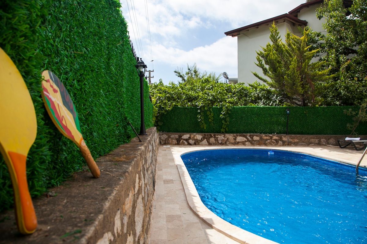 A private swimming pool is framed by green hedges, creating a secluded atmosphere. The pool features clear blue water, and a stone wall surrounds it. Two paddle rackets are placed on the edge, suggesting leisure activities by the water's edge.