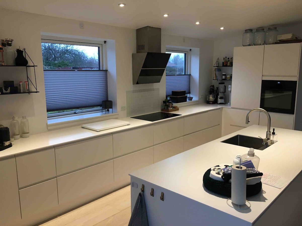 A modern kitchen features sleek white cabinetry and ample counter space. Large windows allow natural light to fill the room, while a stainless steel range hood is positioned above the cooking area. An island with a sink provides additional workspace, complemented by organized items on the counters.