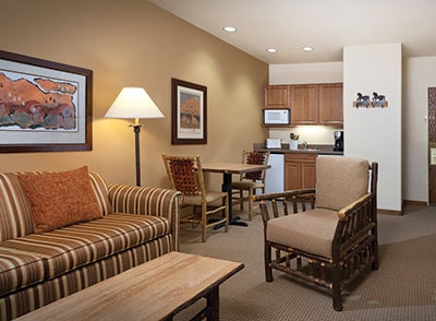 A cozy living area features a striped sofa and a wooden armchair, complemented by a coffee table. Artwork adorns the walls, and a small dining table is set near a partial kitchen with cabinetry and a microwave.