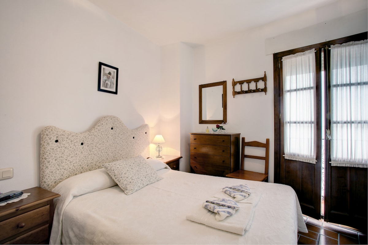 A bright bedroom features a spacious bed with a patterned headboard, accompanied by a bedside lamp providing soft light. A wooden dresser and chair offer additional furnishings, while a large window with sheer curtains allows natural light to fill the room.