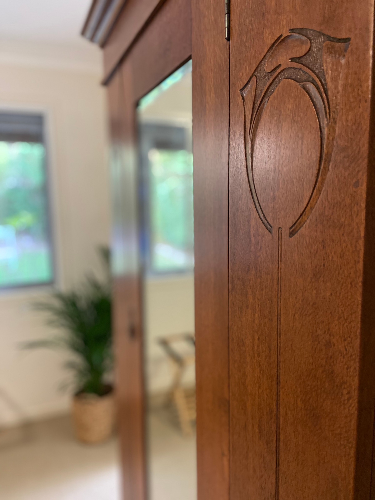 An intricately carved wooden wardrobe is shown, highlighted by its elegant design. The mirror reflects soft natural light, revealing a lush green plant and a subtle seating area in the background, contributing to a calming ambiance.
