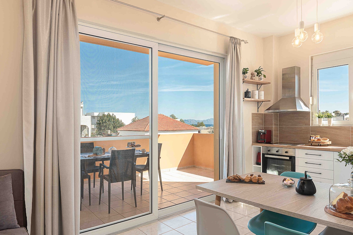A spacious kitchen and dining area showcase a modern design. Large glass doors provide a view of an outdoor dining space with chairs and a table, allowing natural light to fill the room. A counter with an oven and stove is visible, accompanied by a cozy seating arrangement.