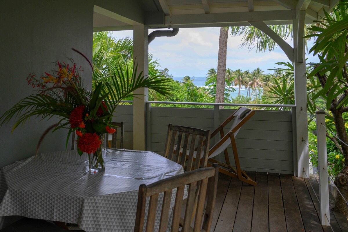 An outdoor terrace features a round dining table with a checkered tablecloth, complemented by wooden chairs. A vase filled with vibrant tropical flowers stands at the center. Lush greenery surrounds the area, with distant views of the sea and palm trees visible.