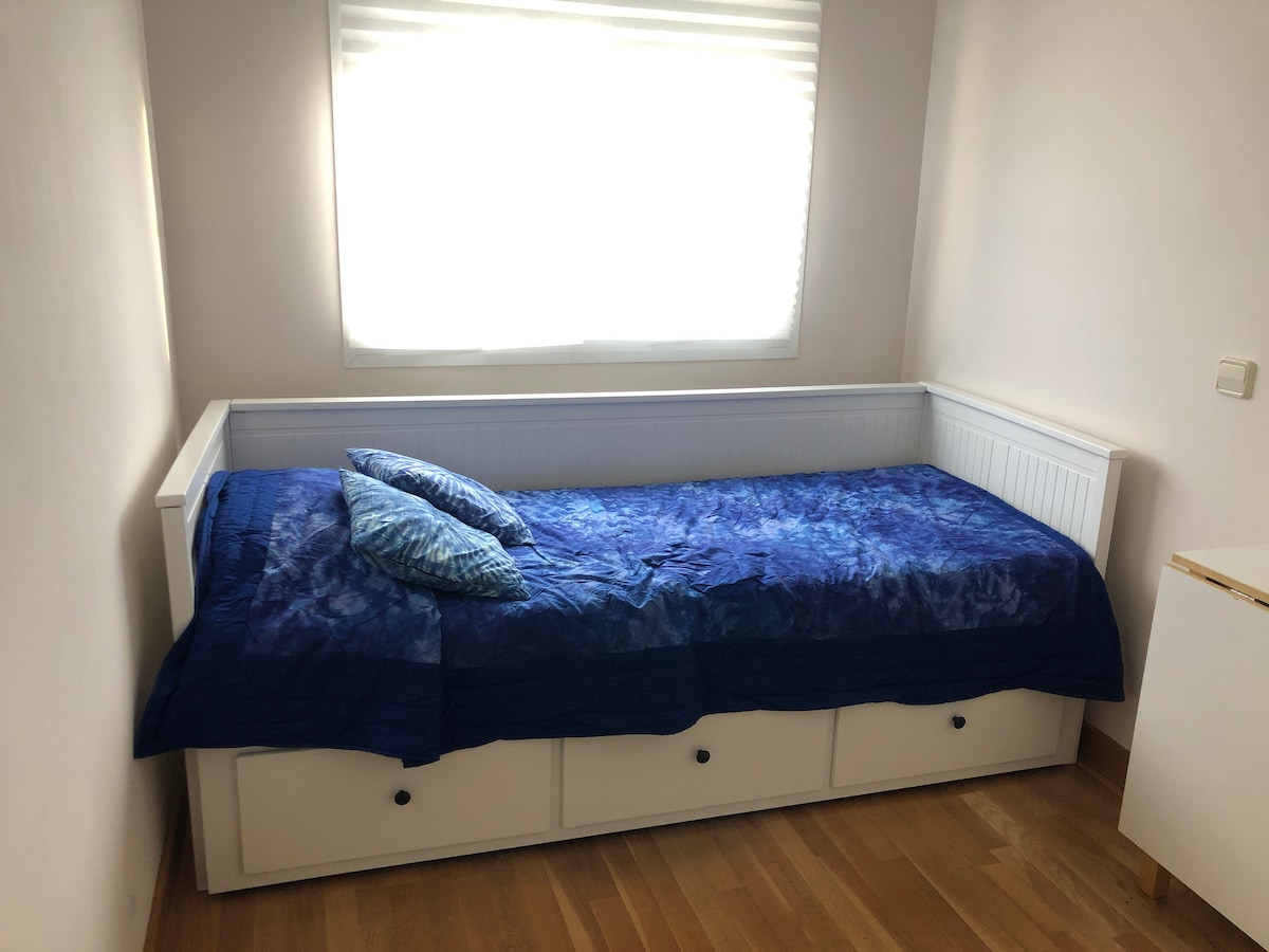 A cozy sleeping area features a white daybed dressed with a blue bedspread and two decorative pillows. Natural light streams in from a nearby window, illuminating the wooden floor and simple white furniture in the room.