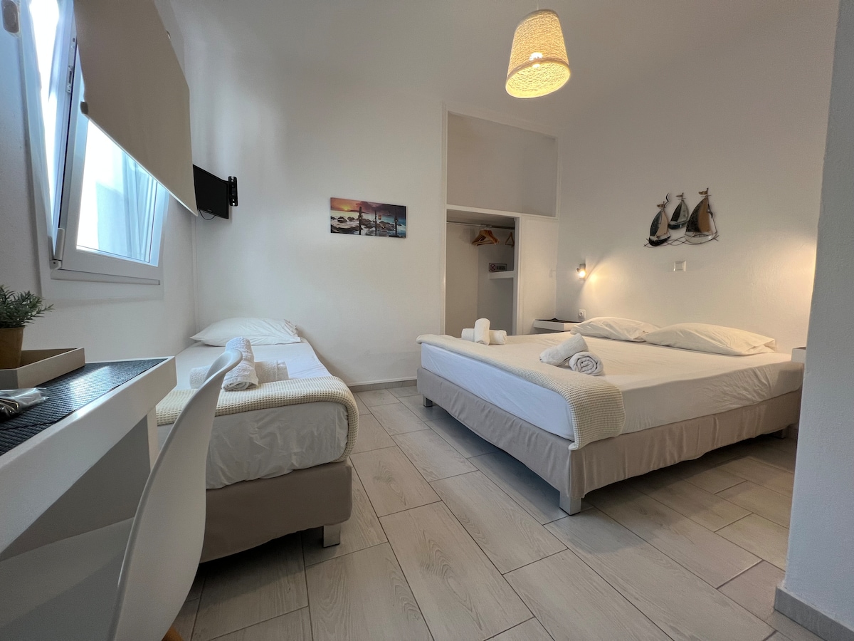 The room features a double bed and a single bed, both dressed in white linens. A small desk and chair are positioned near a bright window. Light-colored tile flooring adds to the airy feel, while decor elements include a wall-mounted television and tasteful artwork.