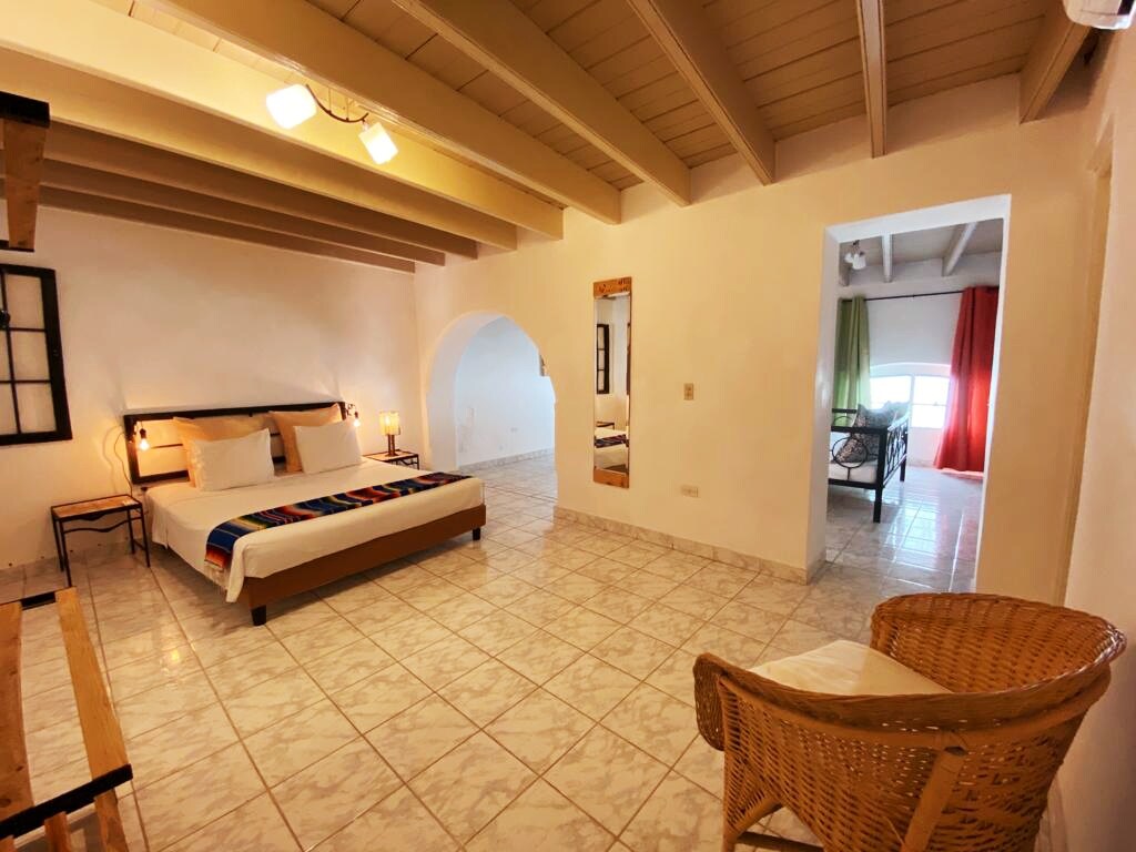 The spacious suite features a bright, open layout with tiled flooring. A comfortable bed is centered against a white wall, while colorful accents can be seen in the textile decor. A wicker chair is positioned beside a wooden bench, with natural light filtering through large windows.