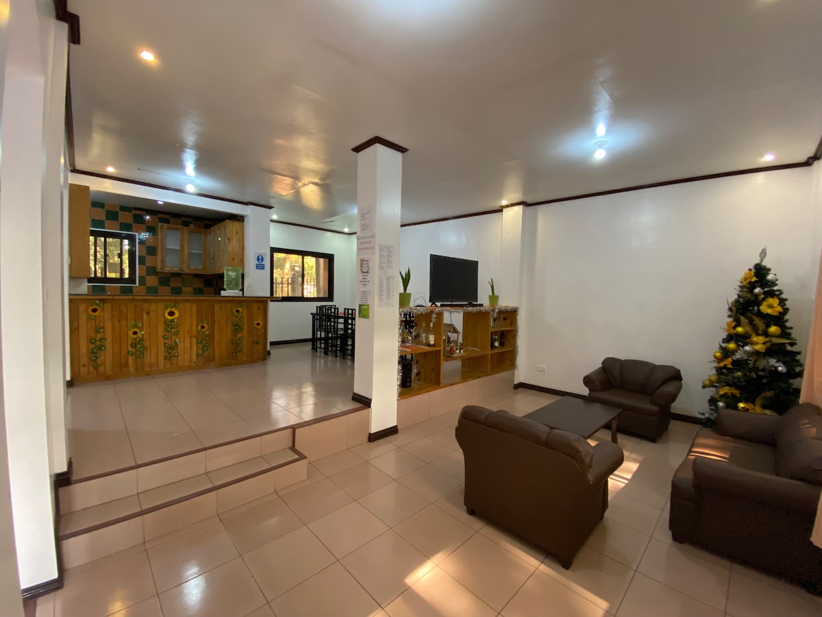 A spacious common area features a welcoming arrangement of brown seating, complemented by a coffee table. The kitchen, visible in the background, includes wood cabinetry and a dining space. A holiday tree adds a festive touch, while natural light filters through large windows.