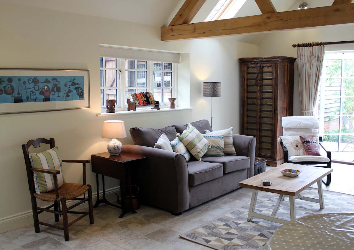 The open-plan sitting area features a comfortable grey sofa adorned with decorative cushions. A wooden table sits in front of the sofa, accompanied by a cozy armchair and a side table. Natural light enters through the French doors, which lead to the garden.