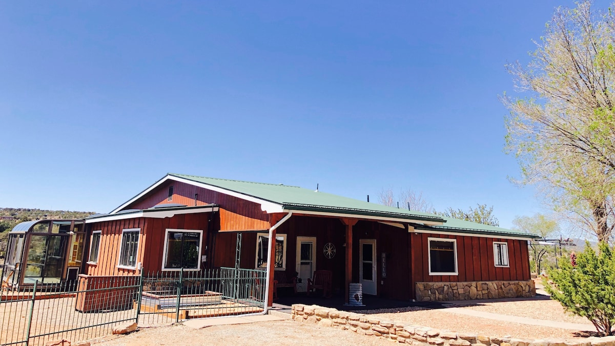 The exterior of the ranch house is shown, featuring a warm wooden façade and a green metal roof. A fenced area is visible in the foreground, with a stone border surrounding the property. Blue skies and distant greenery complement the inviting landscape.