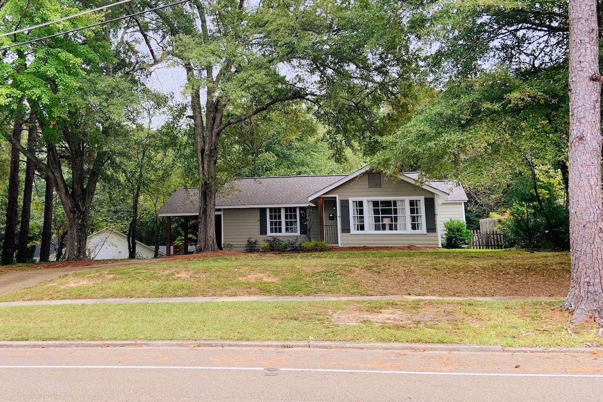 A cozy cottage is set back from the road, surrounded by mature trees and verdant lawns. The front facade features large windows that allow natural light to fill the interior. A pathway leads to a welcoming entry door, enhancing the peaceful neighborhood setting.