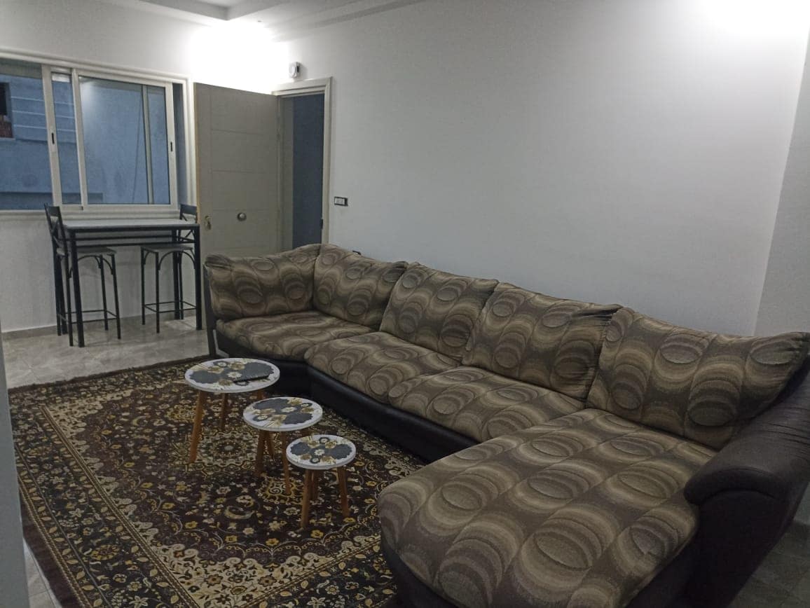 A spacious living area is furnished with a large sectional sofa in neutral tones. Two small coffee tables are positioned on a decorative area rug. A dining set for four is visible near a window, providing natural light to the room.