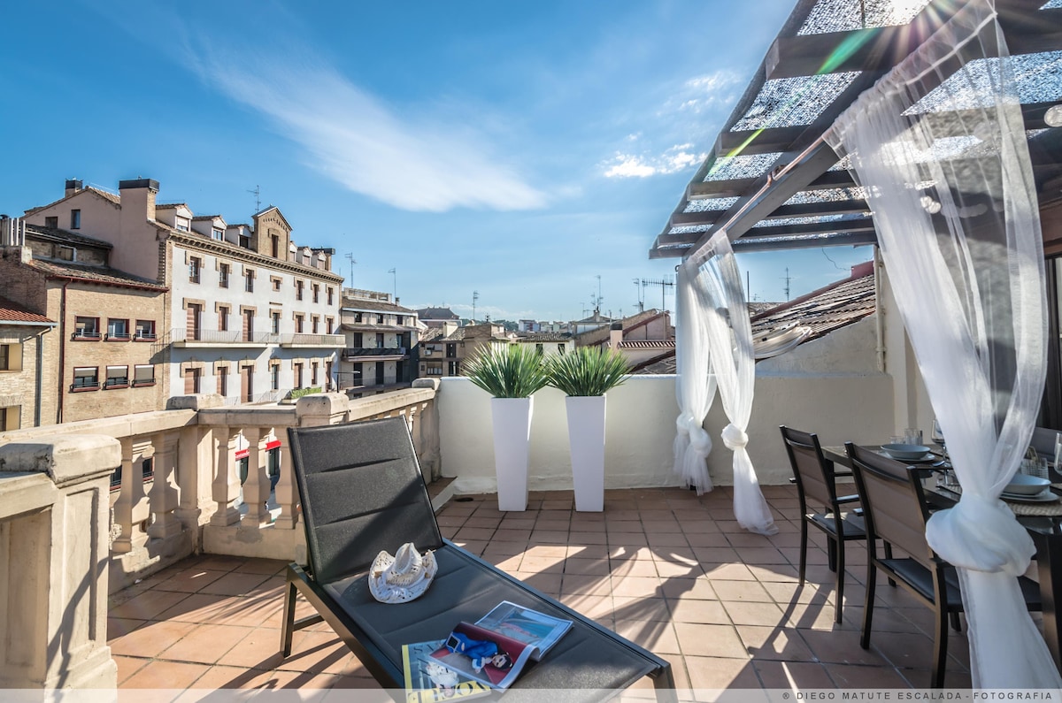 A spacious terrace is showcased, featuring a dining area and a lounge chair. Planters with greenery are positioned at the edges. Sunlight filters through a decorative canopy overhead, creating a bright and open space that overlooks the surrounding buildings and rooftops.