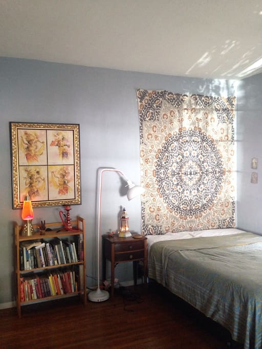 An airy and cozy sleeping space including a queen size bed, dresser, reading lamp, and the mood lighting of a bed side lantern, and lava lamp.