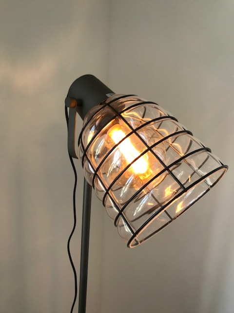 A floor lamp features a metal frame with a protective grid design around the bulb. The warm light emanating from the bulb creates a soft glow, enhancing the ambiance of the space. The lamp is positioned against a simple wall.
