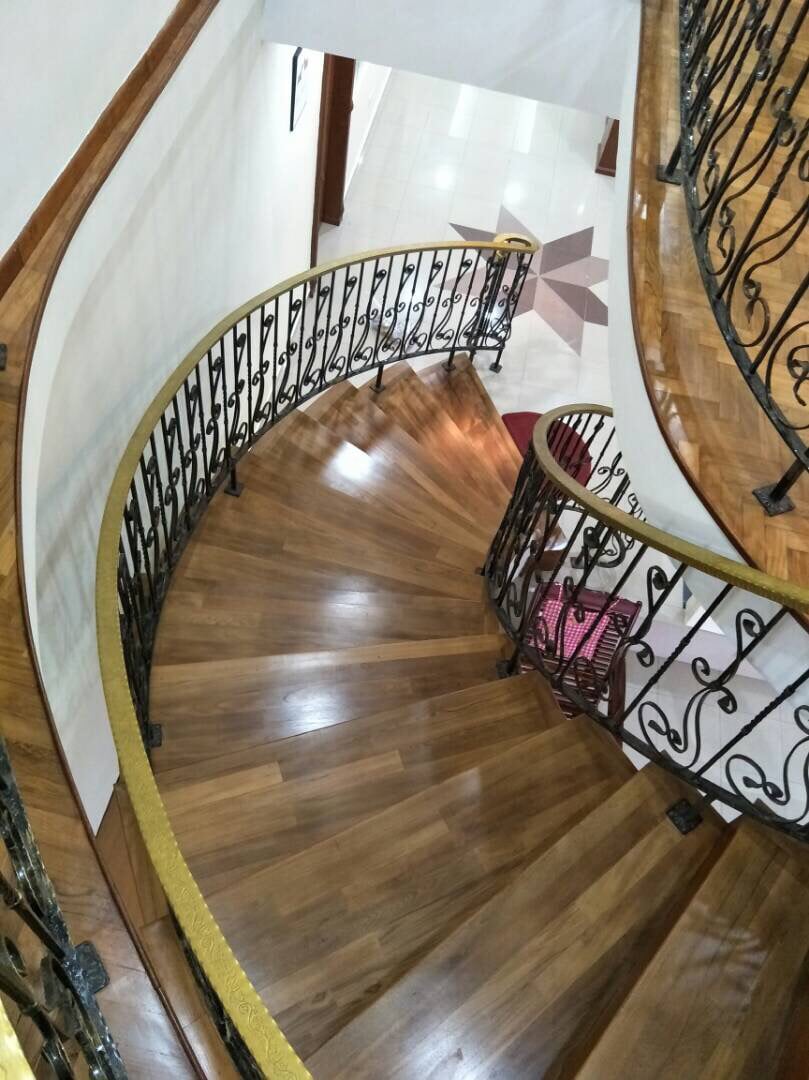 A gracefully winding staircase is depicted, featuring polished wooden steps and a decorative iron railing. Natural light reflects off the surfaces, highlighting the warm wood tones. The staircase leads down to a patterned floor, enriching the overall inviting aesthetic of the space.