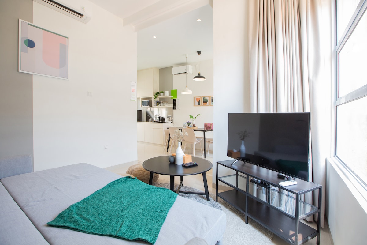 A modern living area showcases a comfortable sofa with a green throw draped over it. A round black coffee table sits in front, complemented by a television on a minimalist stand. Large windows allow natural light to fill the space, creating an inviting atmosphere.