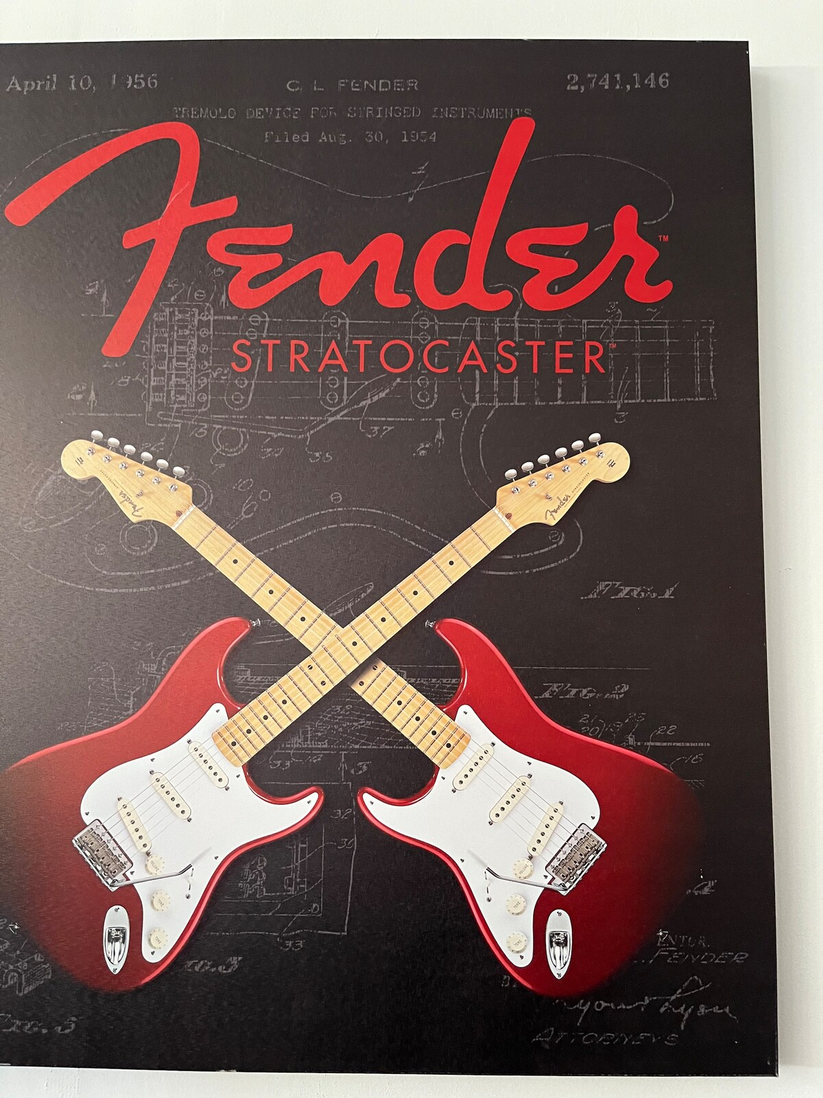 A framed poster featuring two electric guitars, prominently displayed in red and white. The background includes technical drawings and blueprints, providing a contrasting dark backdrop that highlights the instruments. The word 'Fender' is featured in bold red lettering at the top.