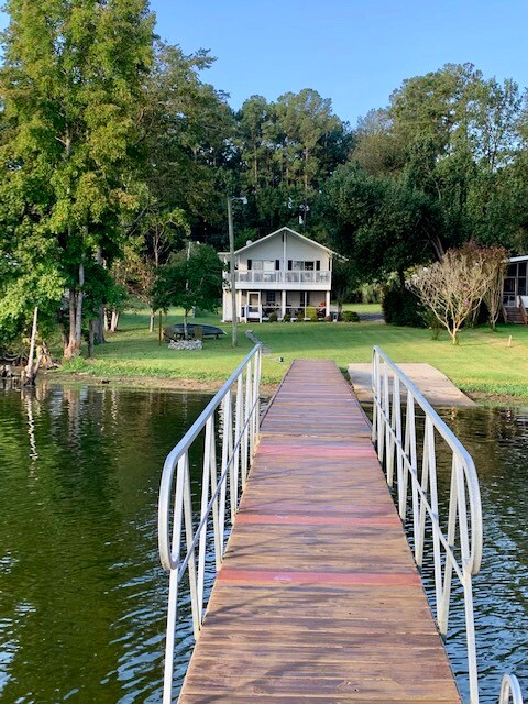 A wooden dock extends towards a two-story lake house, positioned amidst lush greenery. The house features a spacious porch with seating, complemented by the calm lake waters reflecting the surrounding trees. A gentle slope leads from the dock to the well-kept lawn.