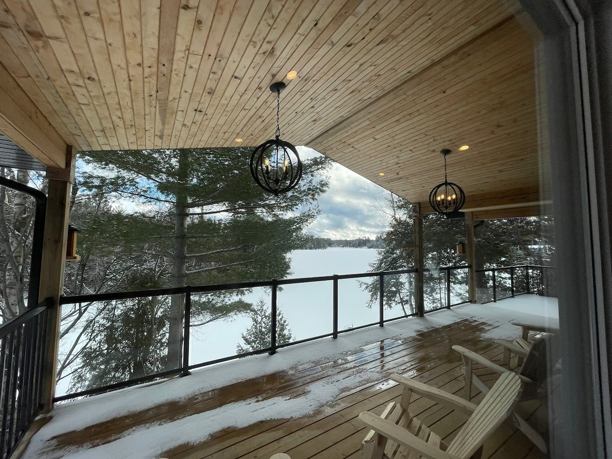 A covered deck features a wooden floor and expansive views of a snow-covered lake. Two suspended light fixtures hang from the wooden ceiling, while a set of white chairs is positioned along the railing. Pine trees can be seen framing the picturesque scene.