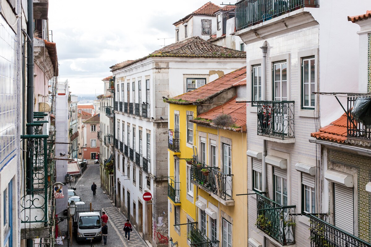 A scenic view of a charming Lisbon street, featuring traditional buildings with decorative balconies and vibrant facades. The narrow, cobblestone road winds through residential areas, showcasing a blend of architectural styles and inviting glimpses of the Lisbon skyline in the distance.
