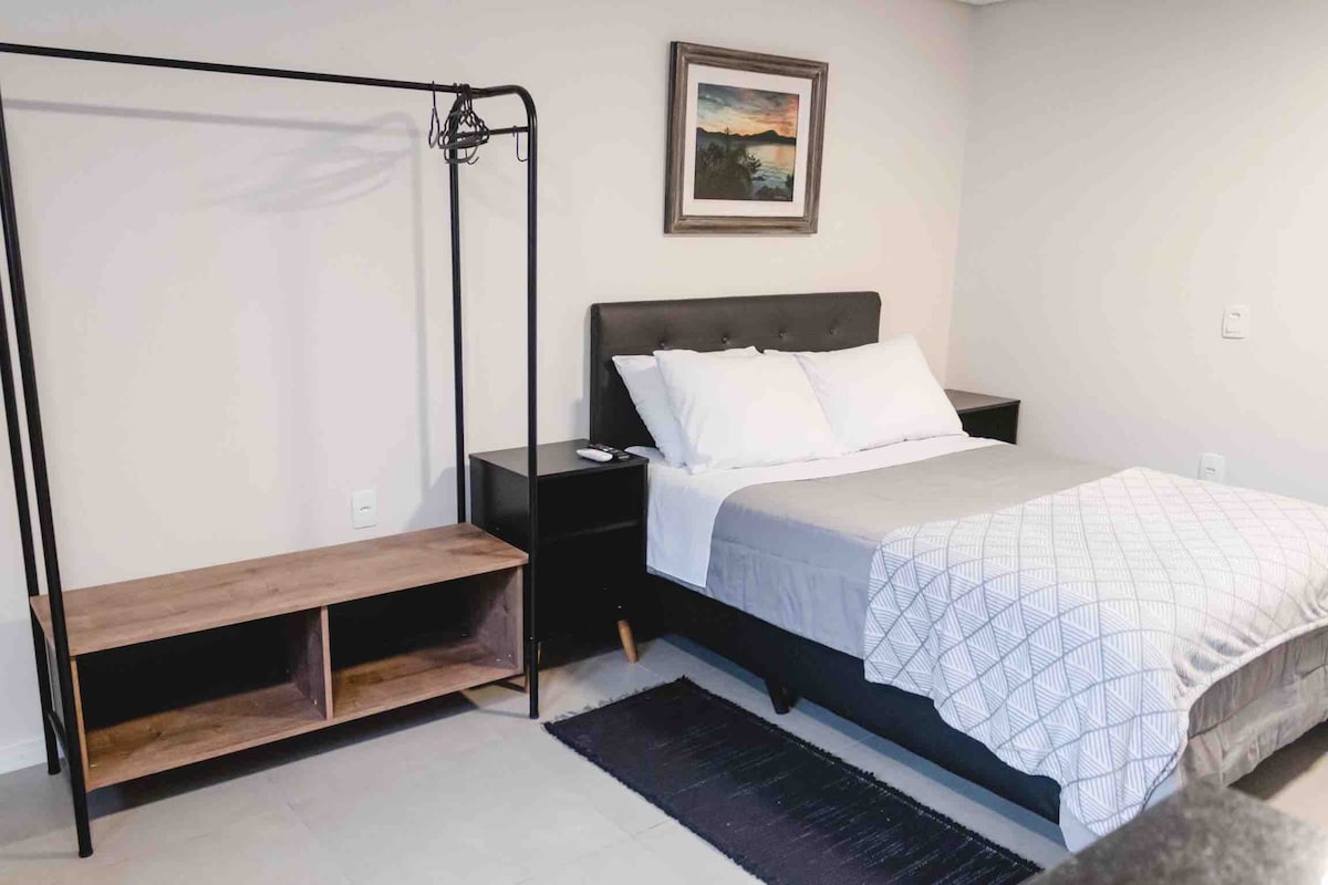 A cozy sleeping area is presented with a spacious bed dressed in white linens. A wall-mounted painting adds a decorative touch. A simple shelving unit and a clothes rack offer functional storage space, while soft lighting illuminates the neutral-toned walls.