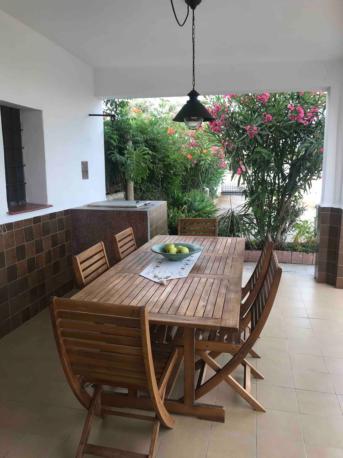 A shaded outdoor dining area features a large wooden table surrounded by folding chairs. A simple decorative centerpiece sits on the table, with lush greenery and blooming flowers visible in the background, creating a pleasant and relaxing environment.