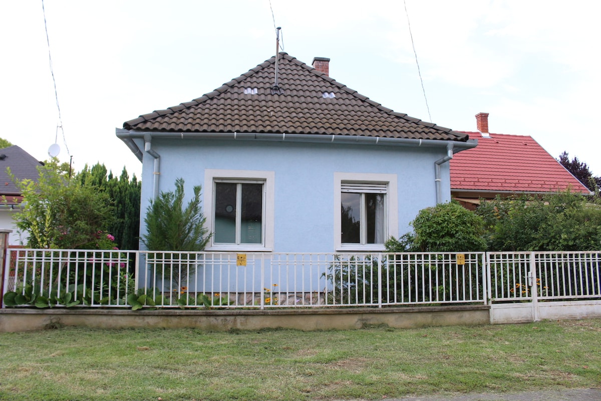 A single-story light blue house with a tiled roof is framed by a white fence and mature greenery. A large window provides a view into the home, while the front yard features a neatly maintained landscape, contributing to the overall welcoming exterior.