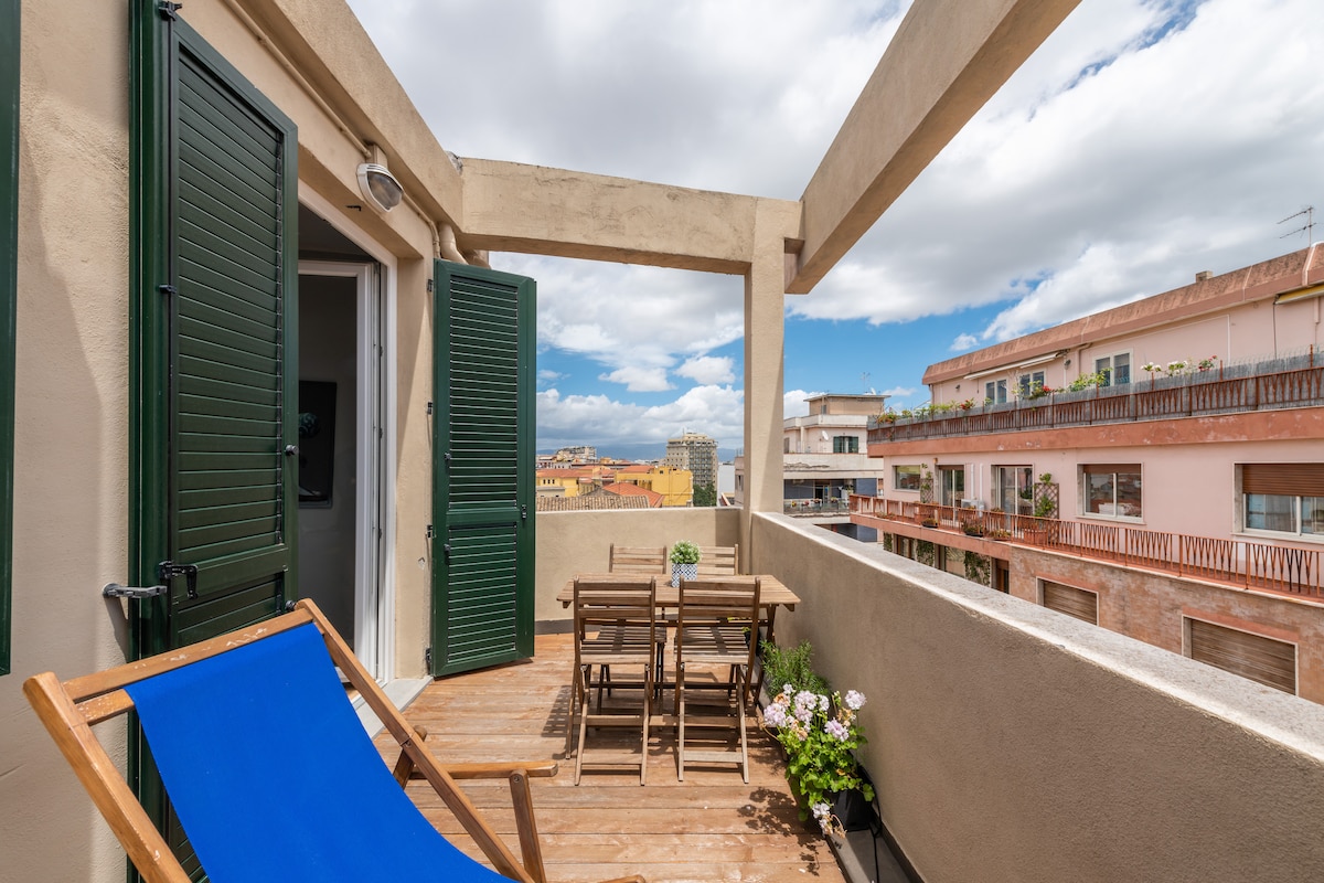 An inviting terrace is featured with a wooden deck, accompanied by a small dining table and chairs. A blue lounge chair is positioned for relaxation. Green shutters frame the entrance, while a view of the city and sea is visible under a partly cloudy sky.