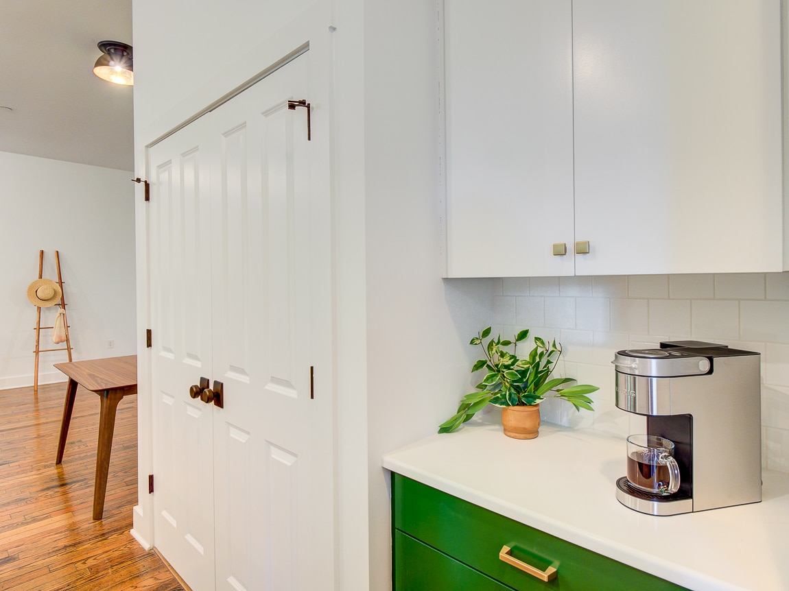A modern kitchen area features sleek white cabinetry and a green lower cabinet. A coffee maker sits atop a countertop next to a small potted plant, with a simple wooden dining table and natural wood flooring visible in the background.